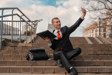 Wall Mural - young man in a black suit and red tie takes a selfie on a smartphone. man with laptop sits on the steps in the city.