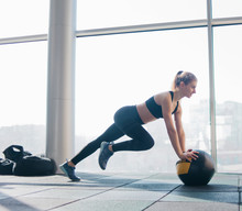 Athletic Attractive Woman Doing Exercise Lifting The Leg Up Leaning On The Medicine Ball Against The Background Of Large Panoramic Window In The Gym. Functional Training
