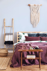Modern and luxury interior of bedroom with design furnitures, stylish macrame, wooden ladder and elegant accessories. Beautiful bed sheets, blankets and pillows. Warm home decor.  Bright and sunny. 