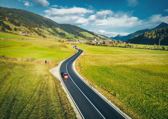 Aerial view of the road in mountain valley at sunset in spring in Dolomites, Italy. Top view of asphalt roadway, car, hills with green meadows, blue sky, trees, buildings. Highway and fields. Nature