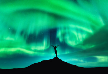 Wall Mural - Aurora borealis and silhouette of a woman with raised up arms on the mountain peak. Lofoten islands, Norway. Aurora and happy girl. Starry sky and polar lights. Night landscape with aurora and people