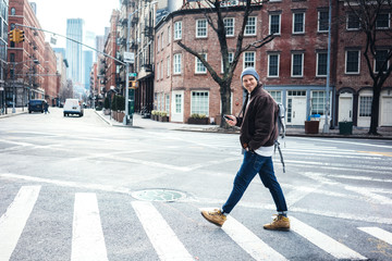 Smiling man wearing hat and jacket walking on the city crossroad with mobile phone in hand