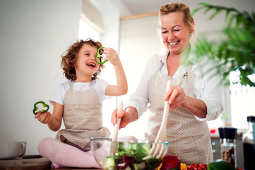 Wall Mural - A portrait of small girl with grandmother at home, preparing vegetable salad.