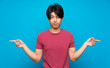 © luismolinero - Asian man with red shirt over isolated blue wall pointing to the laterals having doubts