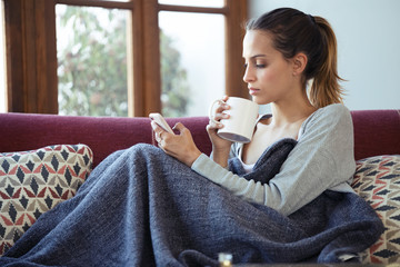 Pretty young woman using her mobile phone while drinking coffee on sofa at home.