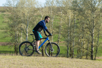 Cyclist in shorts and jersey on a modern carbon hardtail bike with an air suspension fork rides off-road on green hills near the forest