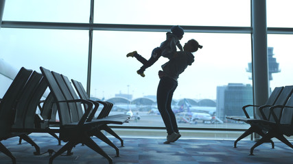 Playful mother raises her son and holds him in arms looking through window in airport.