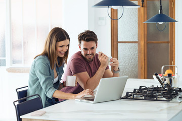 Beautiful lovely young couple using their laptop and having breakfast in the kitchen at home.