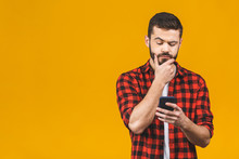 Portrait Of Handsome Young Adult With Dreamy Look, Thinking While Holding Smartphone, Isolated Over Yellow Background. Son Tries To Made Up Message For His Father, Explaining Why He Took Car.