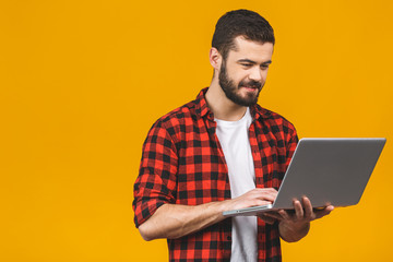 Concentrated on work. Confident young handsome man in casual working on laptop while standing against yellow background.