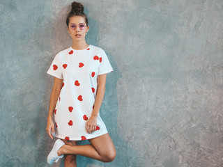 Young beautiful woman looking at camera. Trendy girl in casual summer white dress and sunglasses. Funny and positive female posing near gray wall in studio