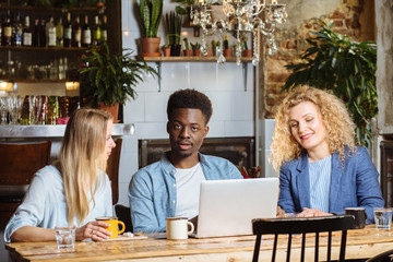 Wall Mural - Multiracial group of three friends having a coffee together. African man and two women at cafe, talking, laughing and enjoying their time. Lifestyle and friendship concepts with real people models