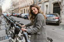 Side View Of Attractive Female In Stylish Coat Cheerfully Smiling And Looking At Camera While Choosing Rental Bicycle On Parking Lot On City Street