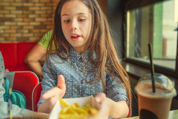 Portrait of a little girl in a fast food restaurant eating fast food, French fries. Happy child eating unhealthy food in restaurant