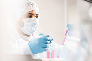 Serious concentrated young Arabian medical scientist in protective workwear and mask comparing liquids in test tubes while working with hazardous substances in laboratory