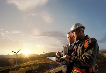 Windmill engineer and businessman planing new ecology project. they standing and looking in tablet. Around wind generators and beautiful landscape