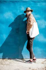 Wall Mural - Portrait of a stylish bearded man dressed in jacket and hat standing with bag on the blue background outdoors