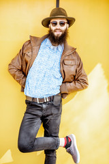 Wall Mural - Full length portrait of a stylish bearded man dressed in jacket and hat on the bright yellow background outdoors