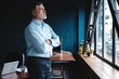© opolja - Mature businessman in a corporate suit standing in office and looking away through large windows optimistically.