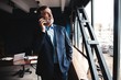 © opolja - Mature businessman talking on a mobile phone standing by the window with view on city.
