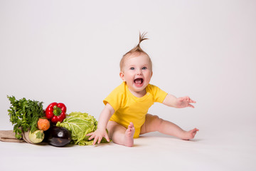 Wall Mural - baby girl smiling in yellow bodysuit on white background with vegetables, baby food concept