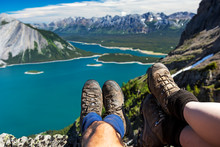 Close-up Of A Couple's Hiking Boots Over A Cliff Overlooking A Colourful Alpine Lake And Mountain Range In The Distance; Kananaskis Country, Alberta, Canada