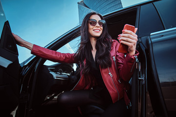 Beautiful smiling women is posing in her new car while chatting on mobile phone. She is wearing red leather jacket and sunglasses.