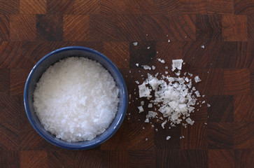 Flake salt lying in a blue bowl and scattered around on a wooden background. Top view image.