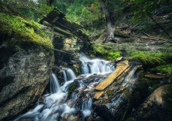 Wall Mural - Stony well in colorful green forest with little waterfall in mountain river at sunset in summer. Landscape with stones in water, building, trees, waterfall and vibrant foliage. Nature. Blurred water