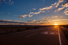 Route 66 Pavement Sign In The Foreground And The Diminishing Perspective Of The Road Leading To A Dramatic Sunset In The Mojave Desert Outside Of Amboy, California.