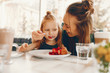 © hetmanstock2 - young and stylish mother with long hair and a green dress sitting with her little cute daughter in the summer cafe and she feeds her daughter with a dessert