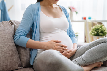 Wall Mural - Focus close up on belly. young elegant asian pregnant woman unrecognized hands touching child in body sitting on couch relax at home in day time in casual wear. chinese girl ready to be mother parent