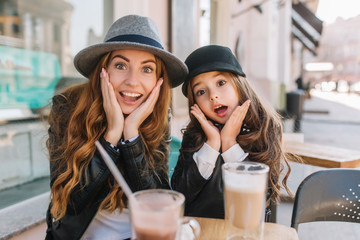 Two amazing girls in trendy hats posing with funny face expression during lunch in street restaurant in sunny day. Young woman with light-brown hair fooling around next to her little daughter