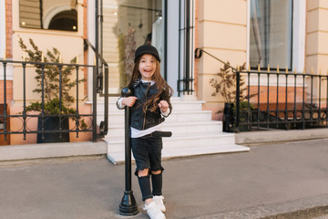 Wall Mural - Full-length portrait of excited kid in grunge outfit funny screaming outside in front of shop. Playful little girl with long brown hair wearing leather clothes fooling around on the street