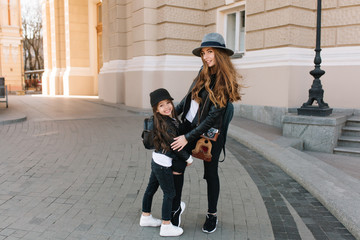 Wall Mural - Excited brunette girl in hat and stylish jacket embracing her mother's leg standing in the middle of street. Joyful good-looking woman with camera spending time in new city with daughter