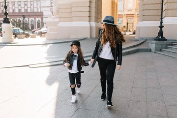 Wall Mural - Slim stylish woman in black jeans holding hand of her cute daughter in leather jacket, walking down the street. Portrait of young mother in felt hat and cheerful long-haired girl having fun in city