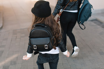 Wall Mural - Portrait of slim graceful mom in black jeans and brunette long-haired daughter in trendy hat playing outside. Playful kid and her shapely mother with backpacks running down the street