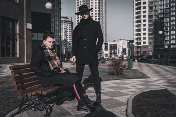 Stylish man street photo shooting in the city. Two businessman posing near bench. Dressed in coat, hat and scarf. Classic style.