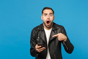 Handsome fun overjoyed excited young unshaven man in black leather jacket white t-shirt using mobile phone isolated on blue wall background studio portrait. People lifestyle concept Mock up copy space