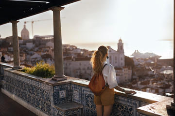 Wall Mural - Blonde woman standing on the balcony and looking at coast view of the southern european city with sea during the sunset, wearing hat, cork bag, safari shorts and white shirt