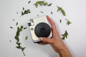 Instant photo camera in the hands of a girl on a background of dry green petals and a camera on a white background. Holding blank instant photo, isolated