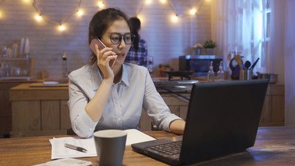 Wall Mural - serious businesswoman talking on smart phone checking mail on netbook laptop late at night. female roommate ready to go bed standing in modern wood kitchen tidy up home waiting for friend finish work