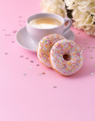 Wall Mural - Mug of hot cappuccino and two donuts with flowers on a pink background.