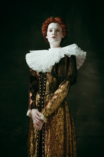 Attention And Serious. Medieval Redhead Young Woman In Golden Vintage Clothing As A Duchess Standing Crossing Hands On Dark Green Background. Concept Of Comparison Of Eras, Modernity And Renaissance.