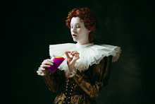 Taste Of Future. Medieval Redhead Young Woman In Golden Vintage Clothing As A Duchess Eating A Fried Potato On Dark Green Background. Concept Of Comparison Of Eras, Modernity And Renaissance.