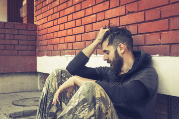 Homeless man drug and alcohol addict sitting alone and depressed on the street leaning against a red brick building wall feeling anxious and lonely in the morning, social documentary concept 
