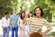 gesture and people concept - happy smiling young woman in striped pullover waving hand over group of friends in summer park background