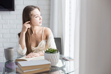 Wall Mural - Beautiful young girl in a nightgown sits in the morning at the glass table, looks out the window and reads a book