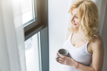 Sticker - Portrait of an attractive blond woman standing near the window with a cup of coffee or tea