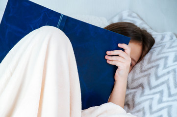 Home portrait of a little girl with long hair sitting in a bed and reading a book